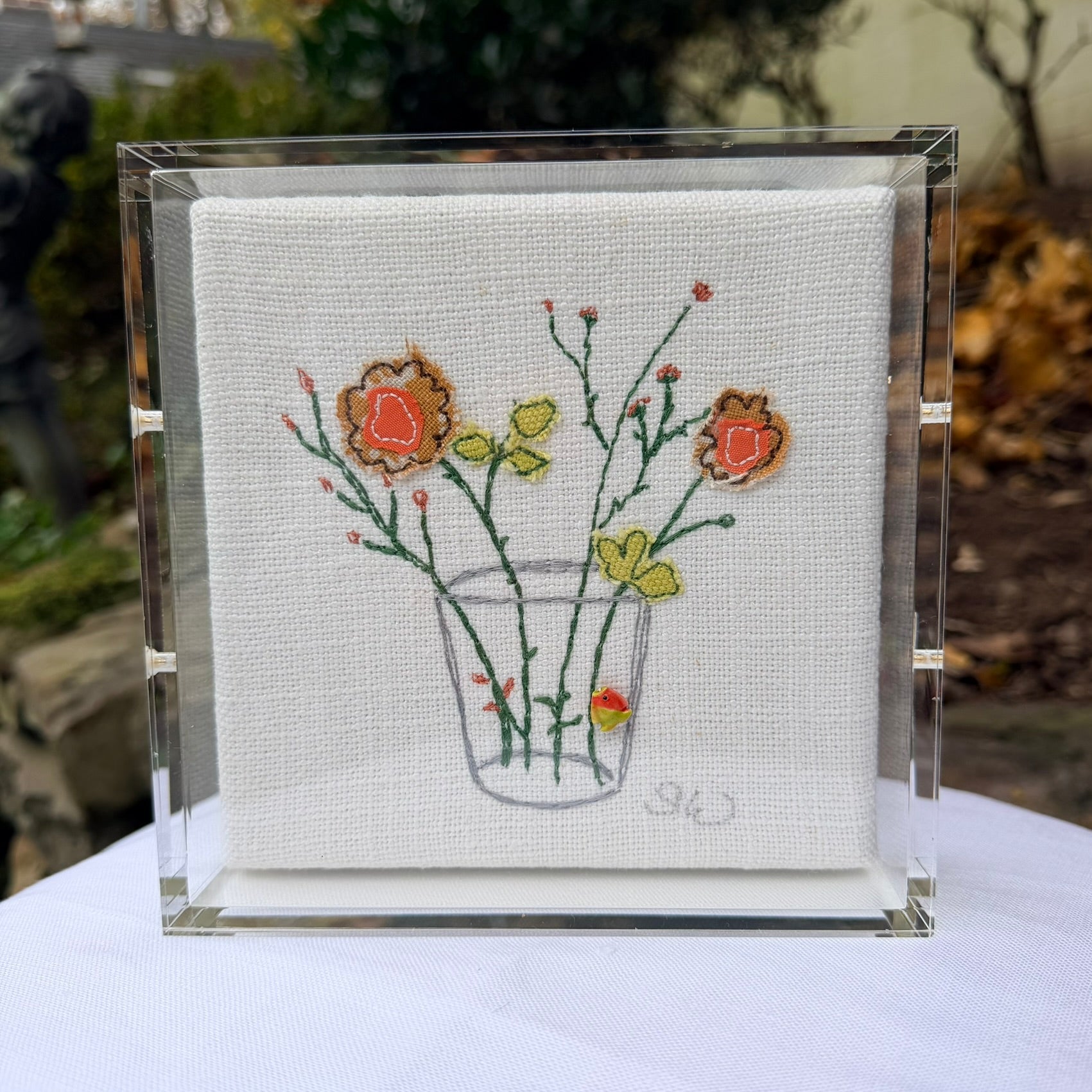 Embroidered artwork of flowers in a glass on a white fabric background, framed in clear acrylic.