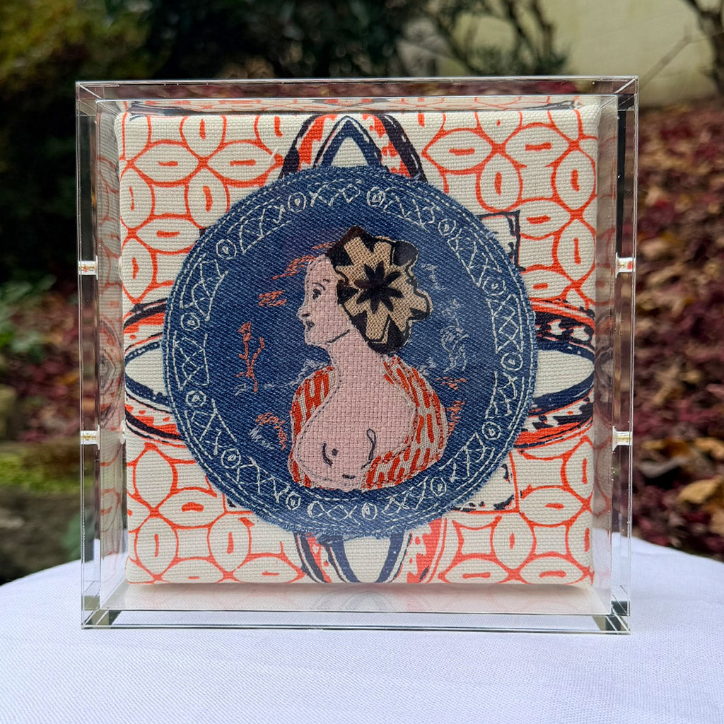 Embroidered patch of a woman&#39;s face in a blue circle on a patterned fabric background, framed in clear acrylic.